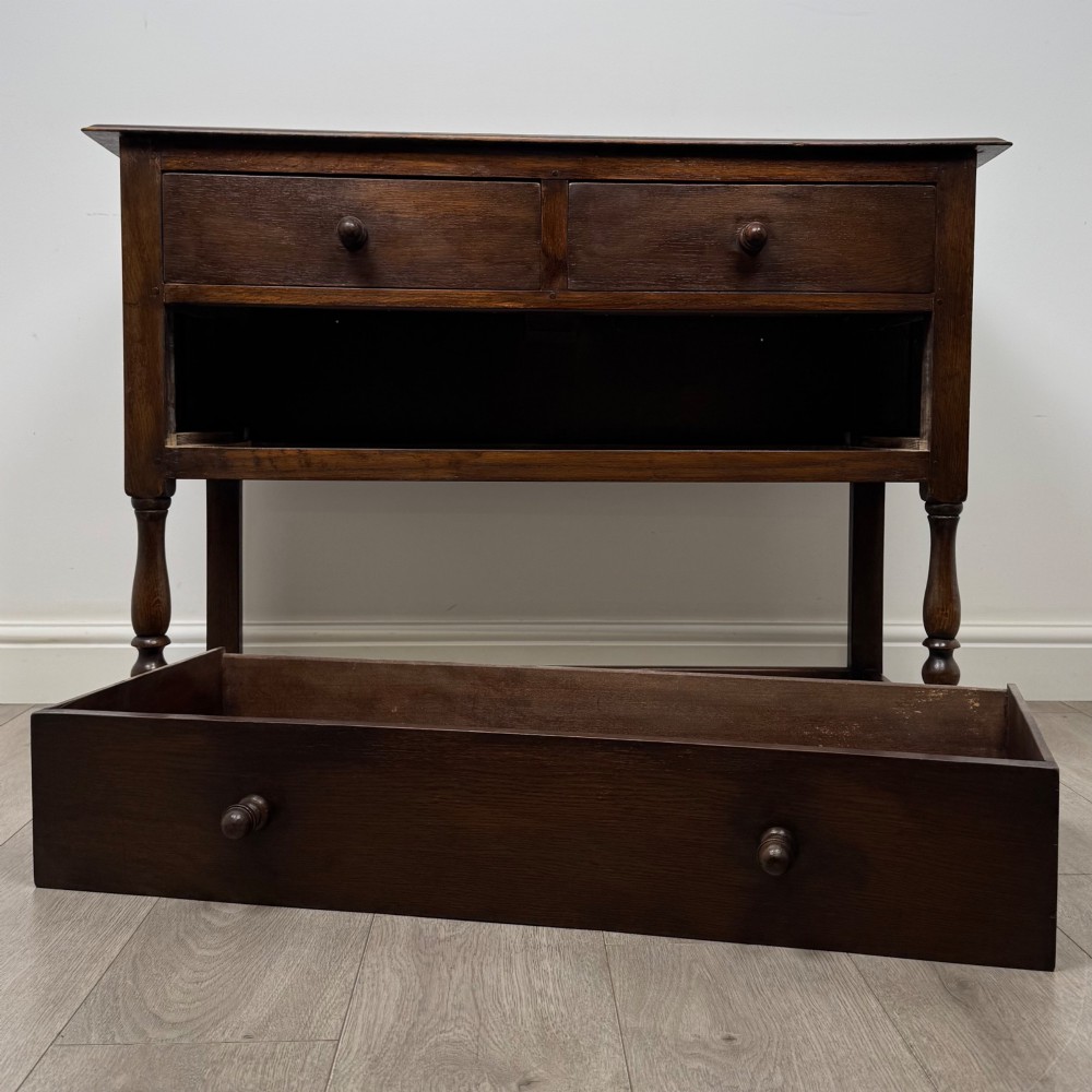 A dark oak chest featuring two top drawers above one wide bottom drawer, resting on turned legs connected by a low stretcher frame. View X