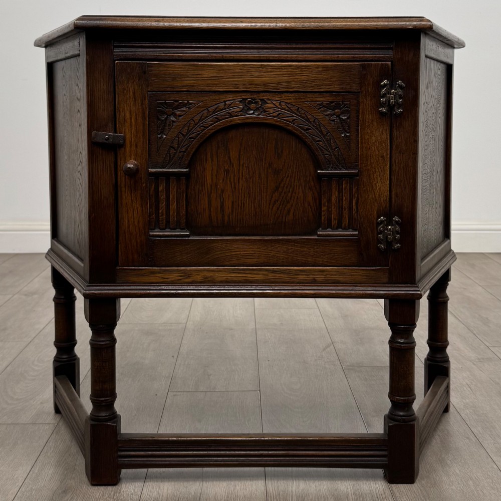 A small dark oak cupboard featuring a single carved panel door, turned legs, a lower stretcher, and decorative brass hinges.