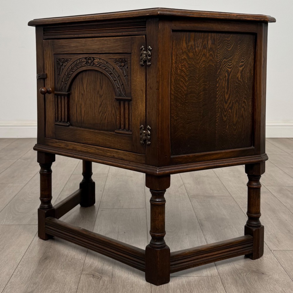 A small dark oak cupboard featuring a single carved panel door, turned legs, a lower stretcher, and decorative brass hinges. View III