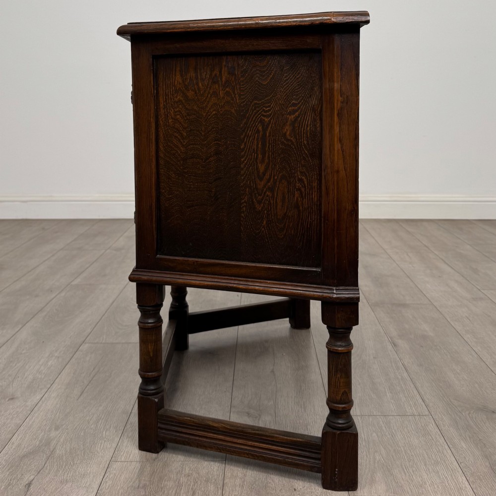 A small dark oak cupboard featuring a single carved panel door, turned legs, a lower stretcher, and decorative brass hinges. View IV