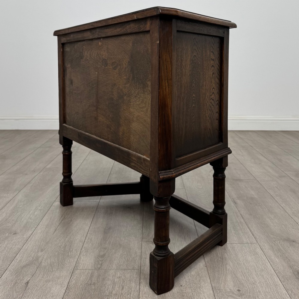 A small dark oak cupboard featuring a single carved panel door, turned legs, a lower stretcher, and decorative brass hinges. View VII