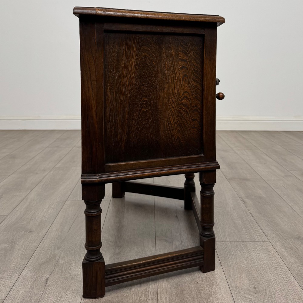 A small dark oak cupboard featuring a single carved panel door, turned legs, a lower stretcher, and decorative brass hinges. View VIII