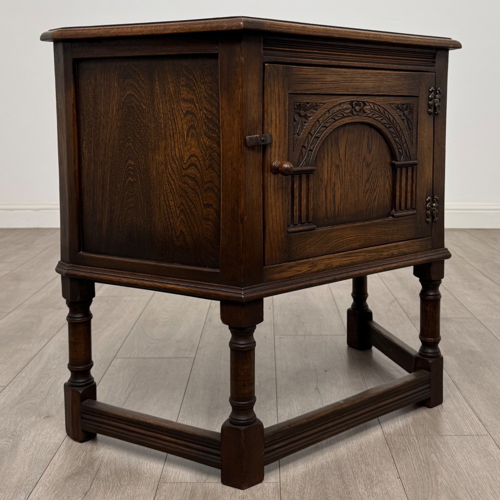 A small dark oak cupboard featuring a single carved panel door, turned legs, a lower stretcher, and decorative brass hinges. View IX