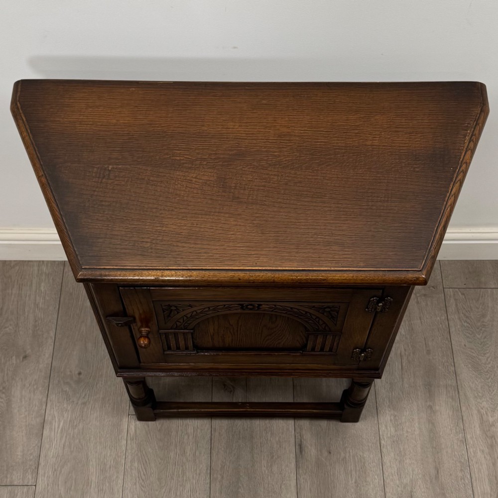 A small dark oak cupboard featuring a single carved panel door, turned legs, a lower stretcher, and decorative brass hinges. View X