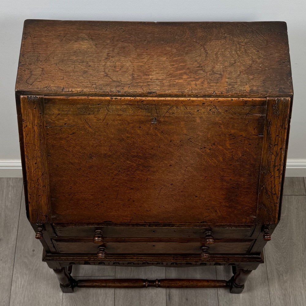 A dark oak antique bureau featuring a slanted drop-front lid above two drawers, supported by turned legs and stretchers. View X