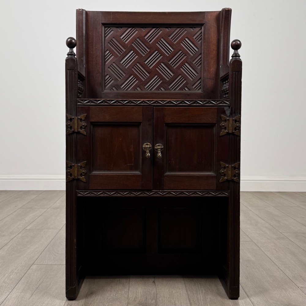 A dark mahogany hall seat cabinet featuring a carved geometric back panel, decorative brass strap hinges, double cupboard doors, and finial corner posts.