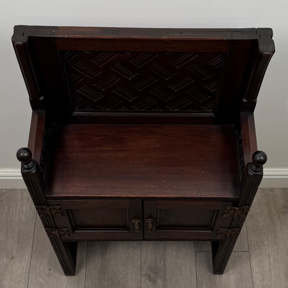 A dark mahogany hall seat cabinet featuring a carved geometric back panel, decorative brass strap hinges, double cupboard doors, and finial corner posts. View X