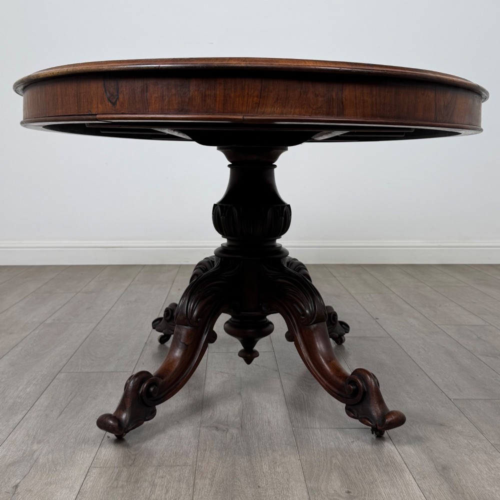 A 19th-century Victorian circular rosewood breakfast table with a carved central pedestal column and four scroll legs with brass castors. View II