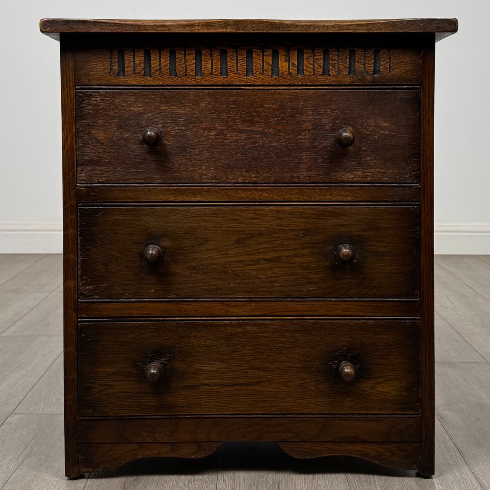 A small early twentieth-century dark oak chest of drawers featuring three drawers with turned wooden knobs and a carved frieze top detail.