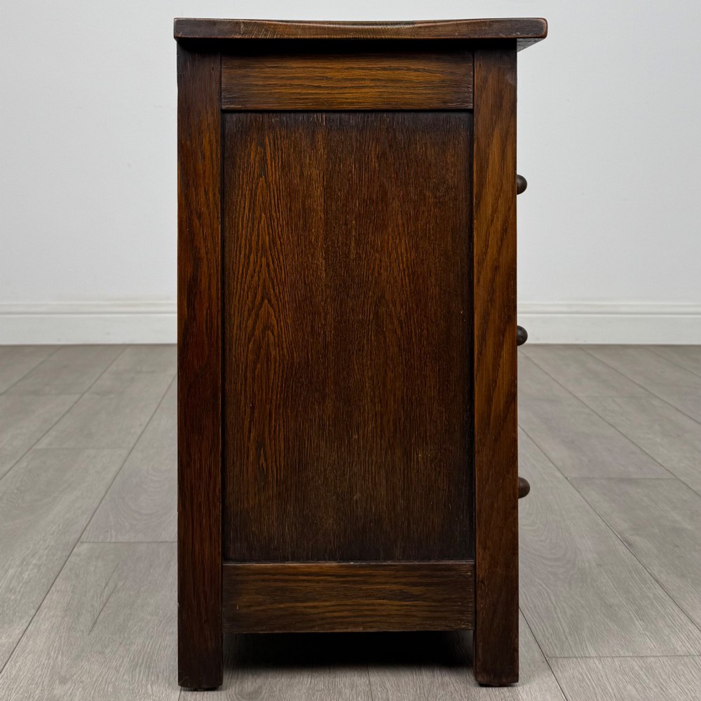 A small early twentieth-century dark oak chest of drawers featuring three drawers with turned wooden knobs and a carved frieze top detail. View VII
