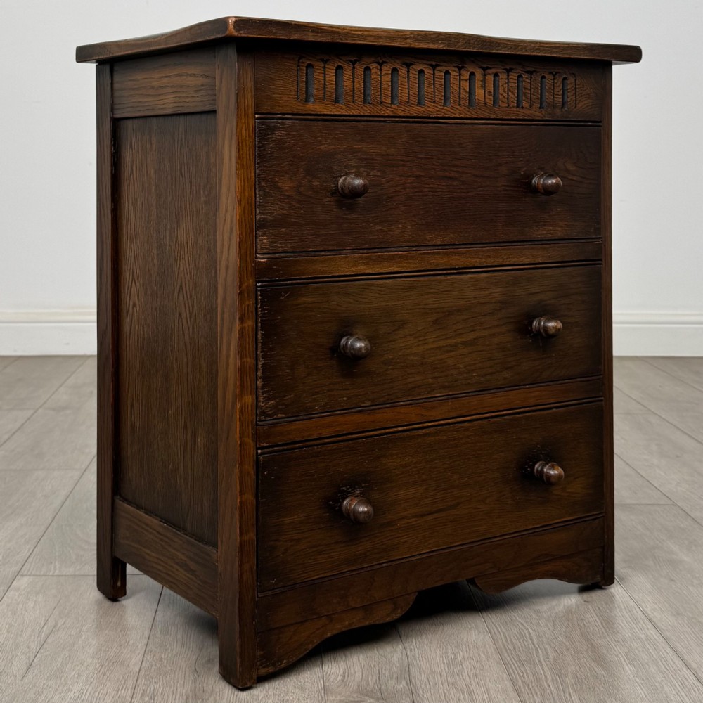 A small early twentieth-century dark oak chest of drawers featuring three drawers with turned wooden knobs and a carved frieze top detail. View VIII