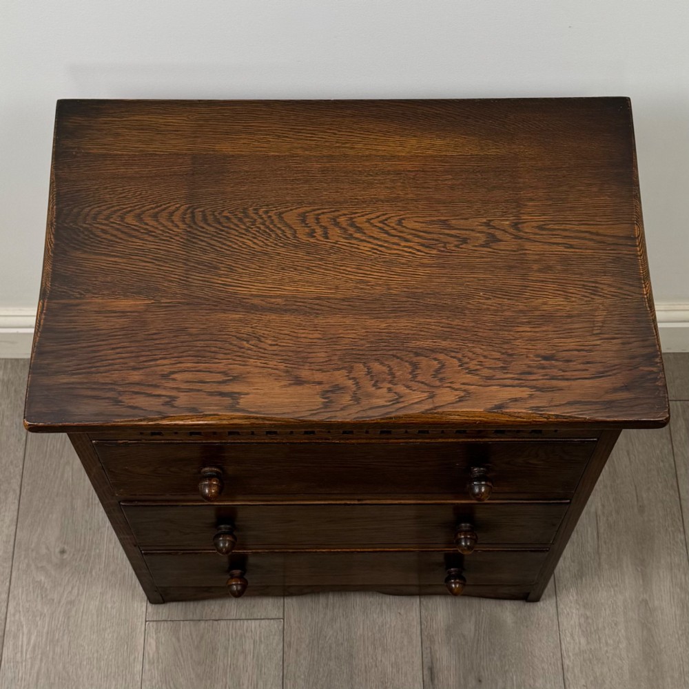 A small early twentieth-century dark oak chest of drawers featuring three drawers with turned wooden knobs and a carved frieze top detail. View IX