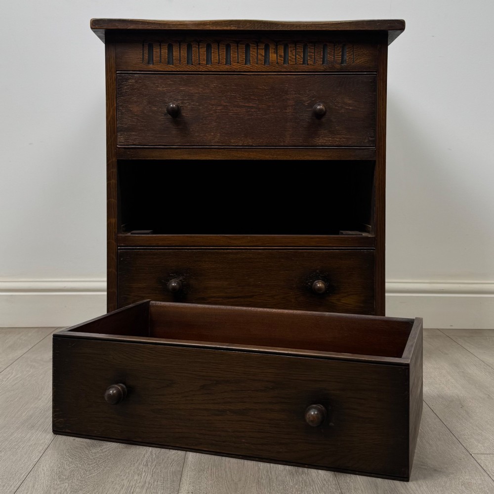 A small early twentieth-century dark oak chest of drawers featuring three drawers with turned wooden knobs and a carved frieze top detail. View X
