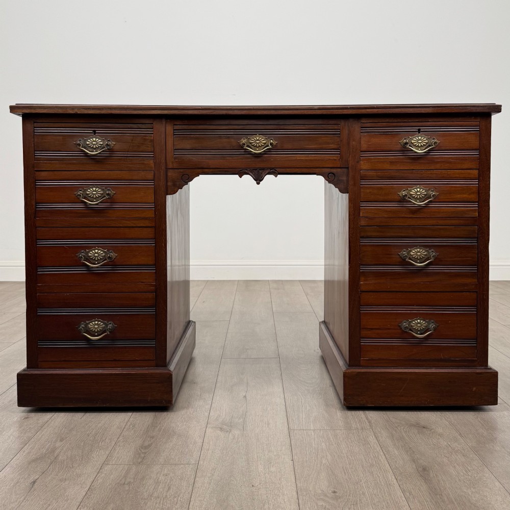 An antique Edwardian walnut pedestal desk featuring nine drawers with reeded panel details, brass drop handles, and a carved kneehole arch.