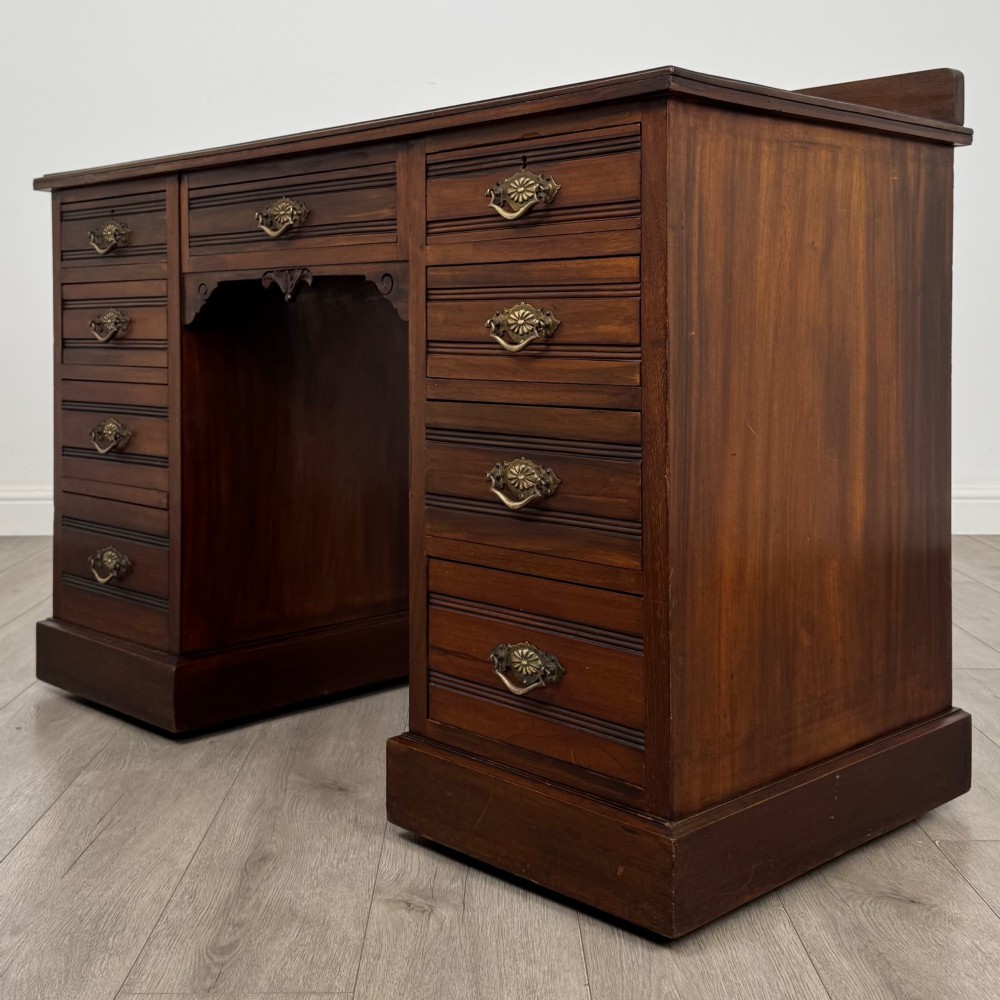 An antique Edwardian walnut pedestal desk featuring nine drawers with reeded panel details, brass drop handles, and a carved kneehole arch. View II