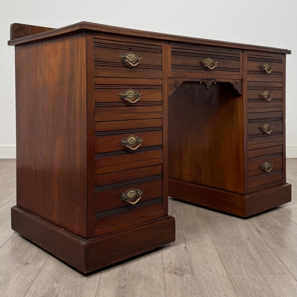 An antique Edwardian walnut pedestal desk featuring nine drawers with reeded panel details, brass drop handles, and a carved kneehole arch. View VIII