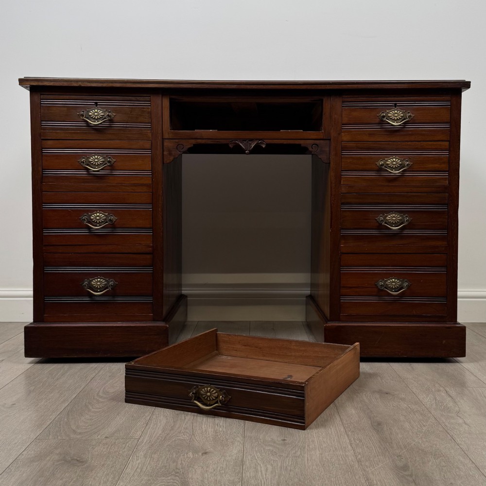 An antique Edwardian walnut pedestal desk featuring nine drawers with reeded panel details, brass drop handles, and a carved kneehole arch. View X