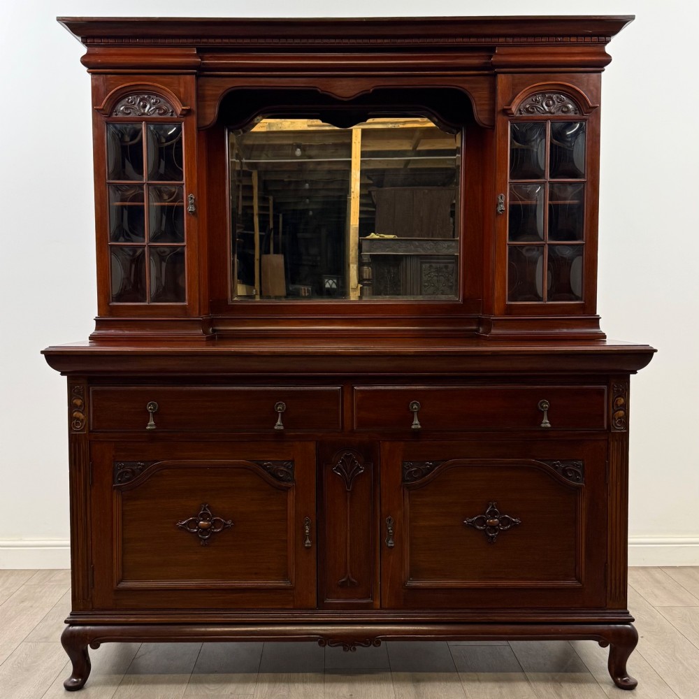 An Arts and Crafts dark mahogany sideboard featuring an upper mirrored superstructure flanked by leaded glass cabinet doors above lower drawers and carved cupboards.