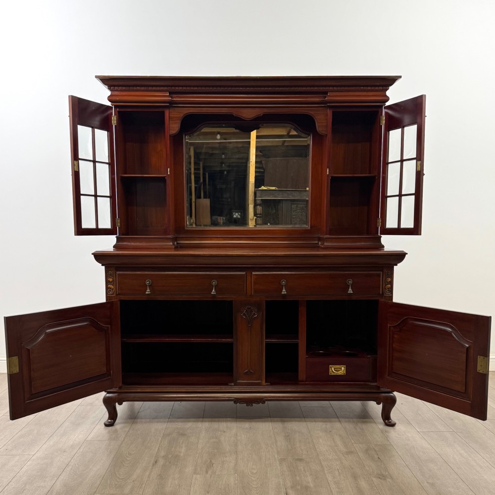 An Arts and Crafts dark mahogany sideboard featuring an upper mirrored superstructure flanked by leaded glass cabinet doors above lower drawers and carved cupboards. View II