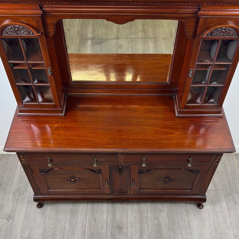 An Arts and Crafts dark mahogany sideboard featuring an upper mirrored superstructure flanked by leaded glass cabinet doors above lower drawers and carved cupboards. View X