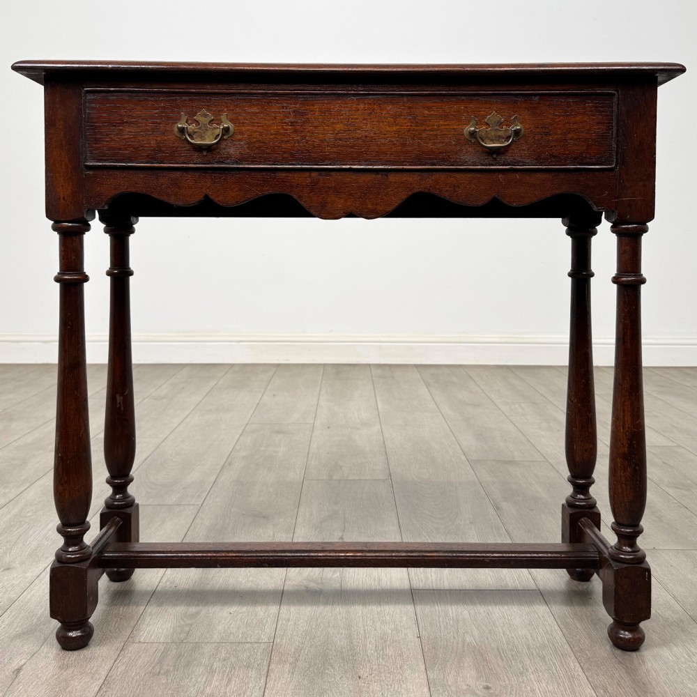 A dark oak side table featuring a single wide drawer with brass drop handles, a scalloped apron, turned legs, and lower stretchers.