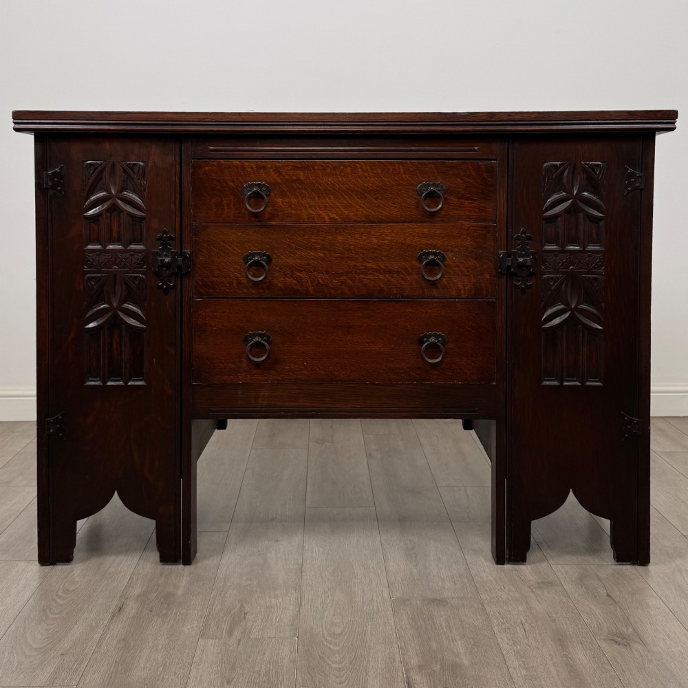 A dark oak Gothic Revival sideboard featuring three central drawers with metal drop ring handles, flanked by two carved tracery panel doors with decorative iron latches.