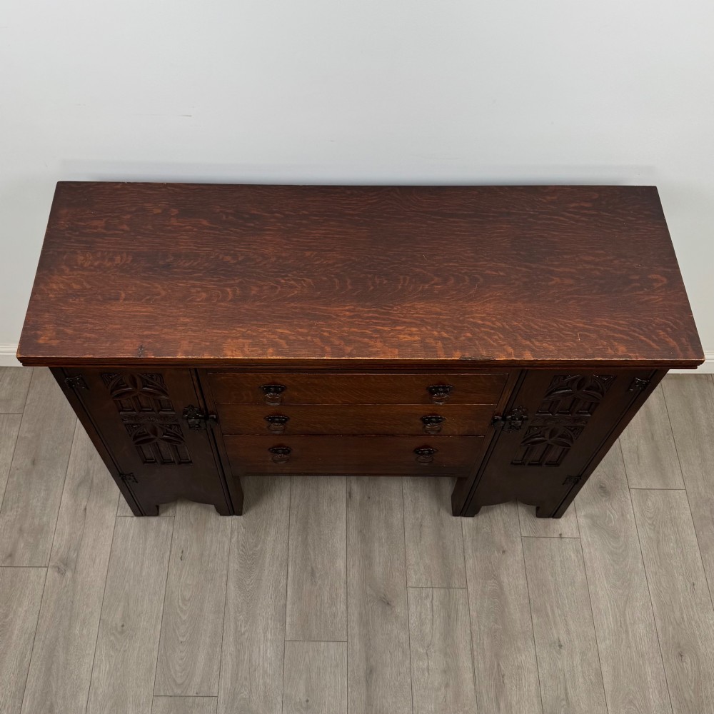 A dark oak Gothic Revival sideboard featuring three central drawers with metal drop ring handles, flanked by two carved tracery panel doors with decorative iron latches. View X