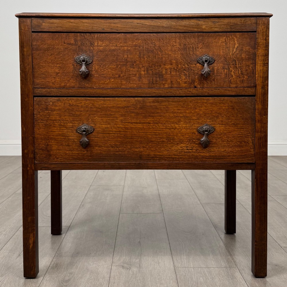 A two-drawer golden oak chest of drawers featuring decorative metal drop handles and straight square legs.