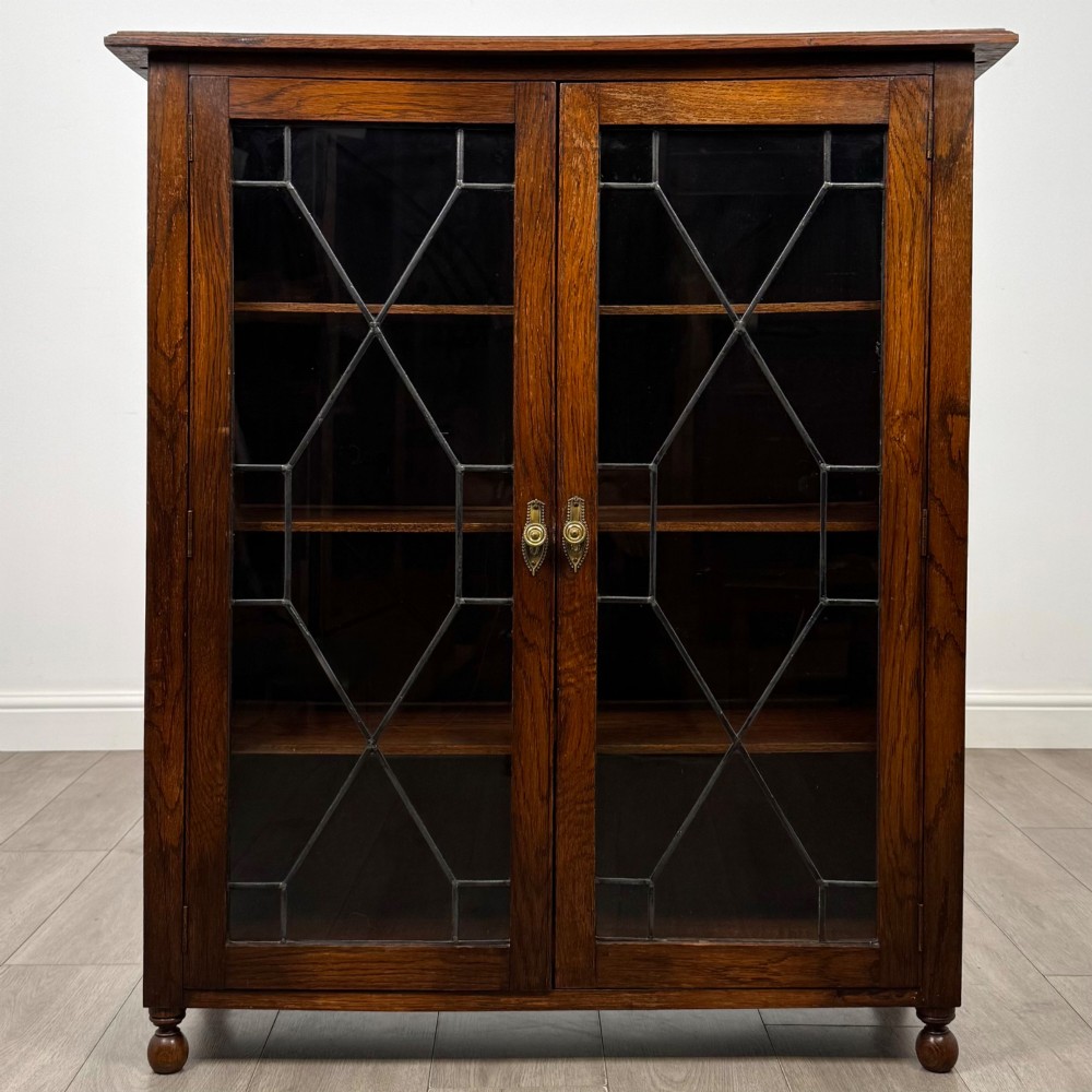 An Edwardian warm brown oak display cabinet featuring twin leaded glass doors with a geometric pattern, interior wooden shelves, and rounded bun feet.