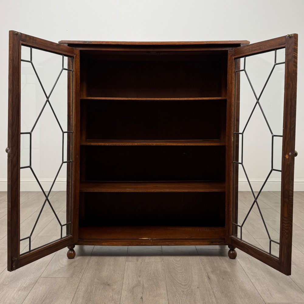 An Edwardian warm brown oak display cabinet featuring twin leaded glass doors with a geometric pattern, interior wooden shelves, and rounded bun feet. View II