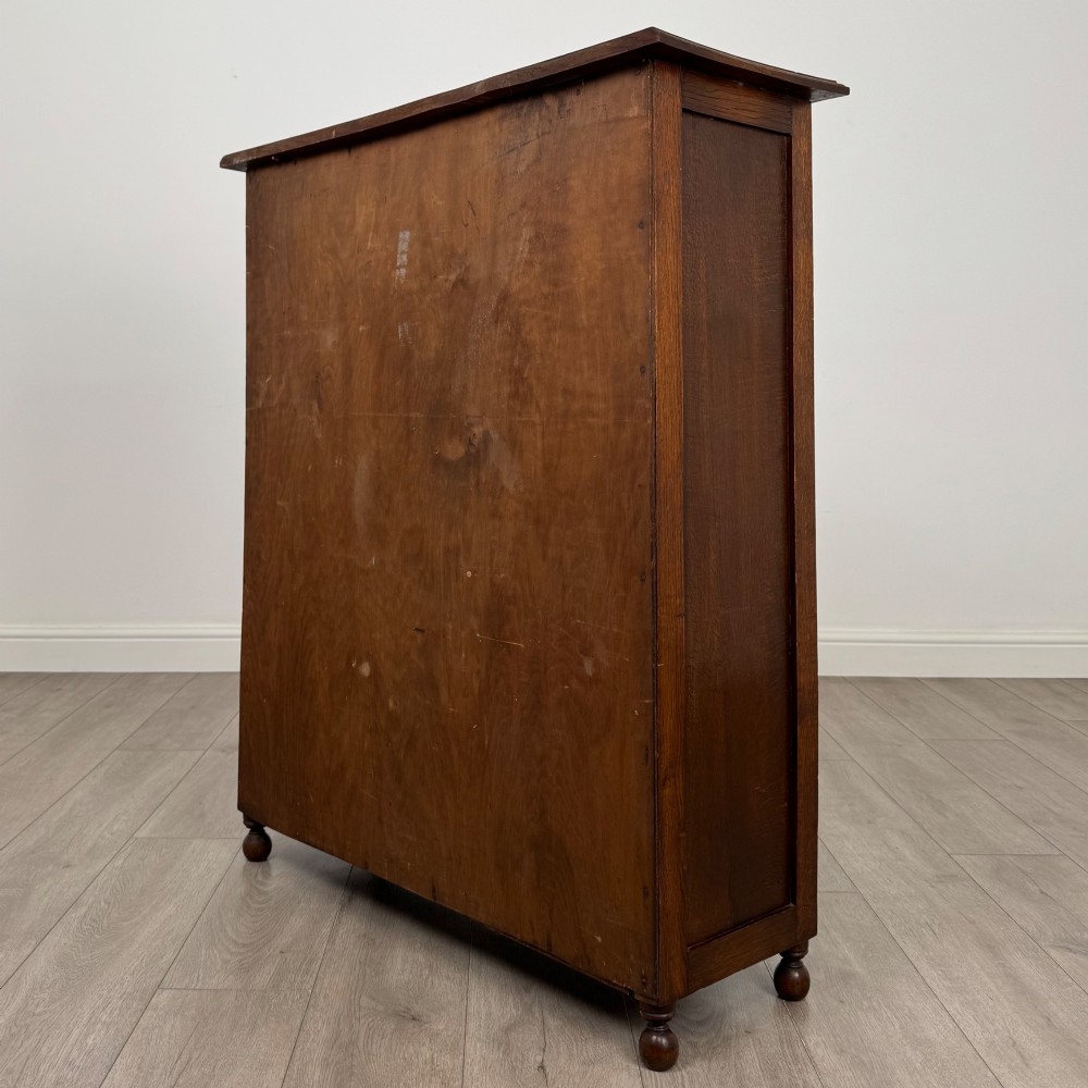 An Edwardian warm brown oak display cabinet featuring twin leaded glass doors with a geometric pattern, interior wooden shelves, and rounded bun feet. View VII