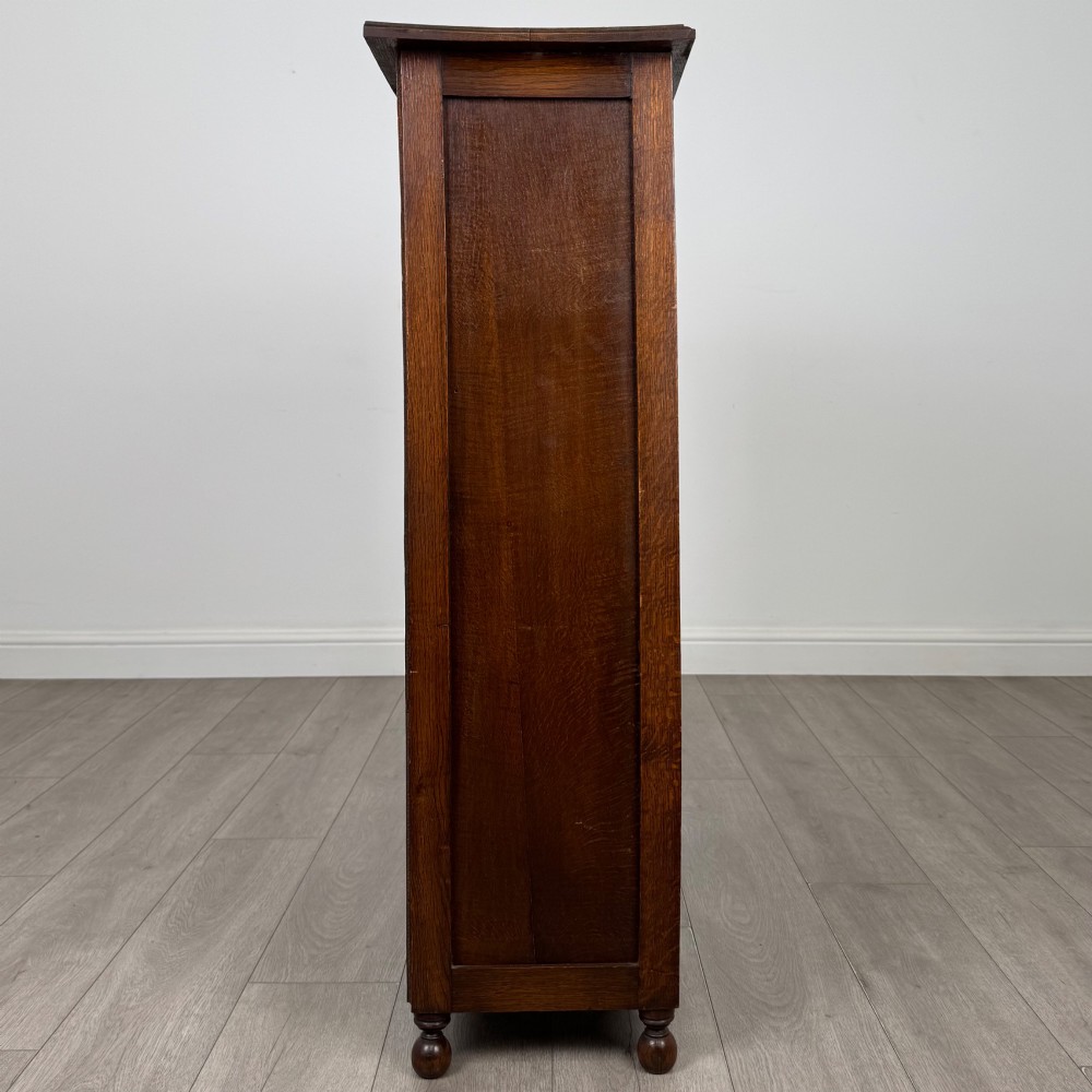 An Edwardian warm brown oak display cabinet featuring twin leaded glass doors with a geometric pattern, interior wooden shelves, and rounded bun feet. View VIII