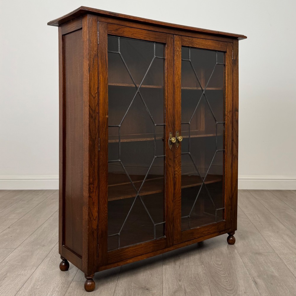 An Edwardian warm brown oak display cabinet featuring twin leaded glass doors with a geometric pattern, interior wooden shelves, and rounded bun feet. View IX