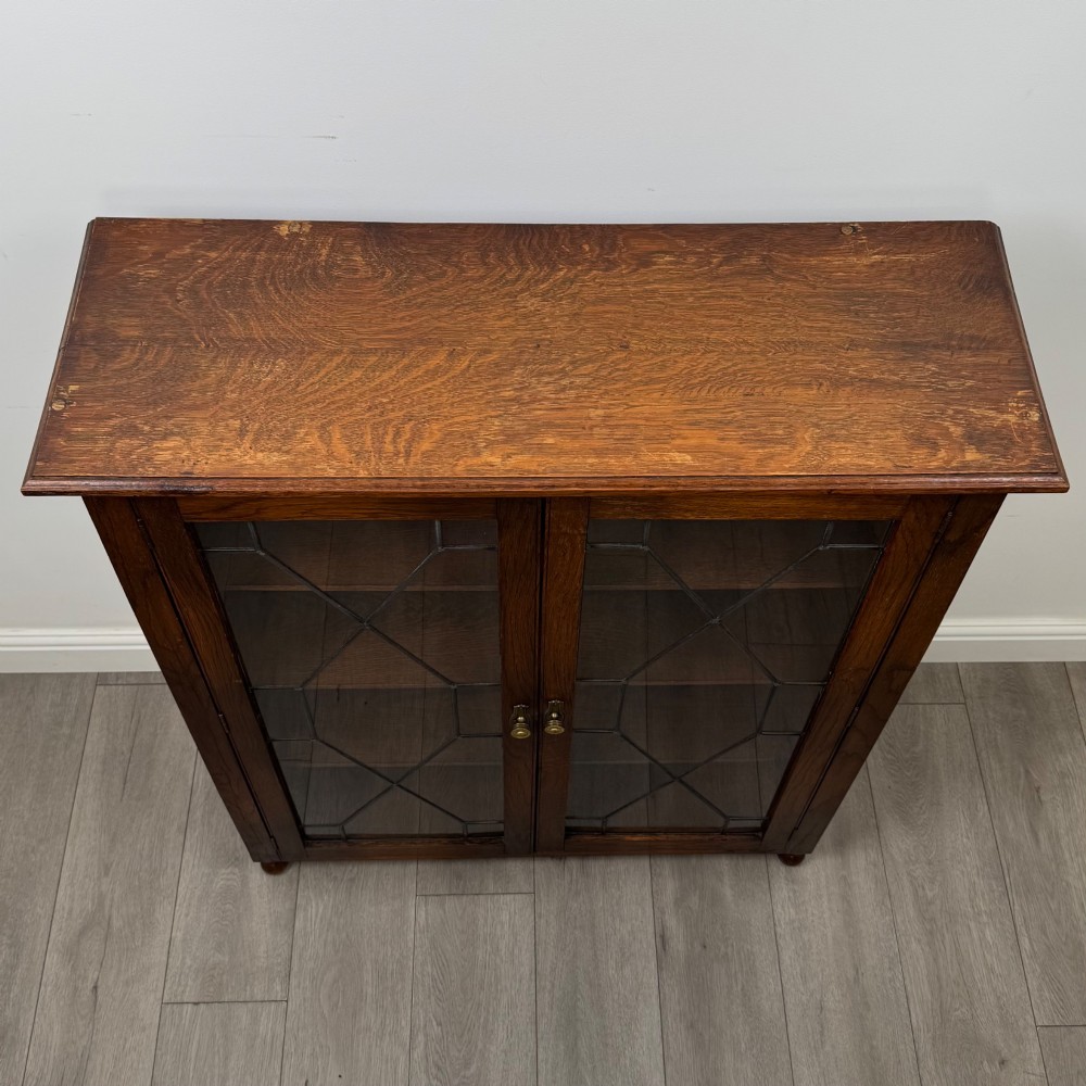 An Edwardian warm brown oak display cabinet featuring twin leaded glass doors with a geometric pattern, interior wooden shelves, and rounded bun feet. View X