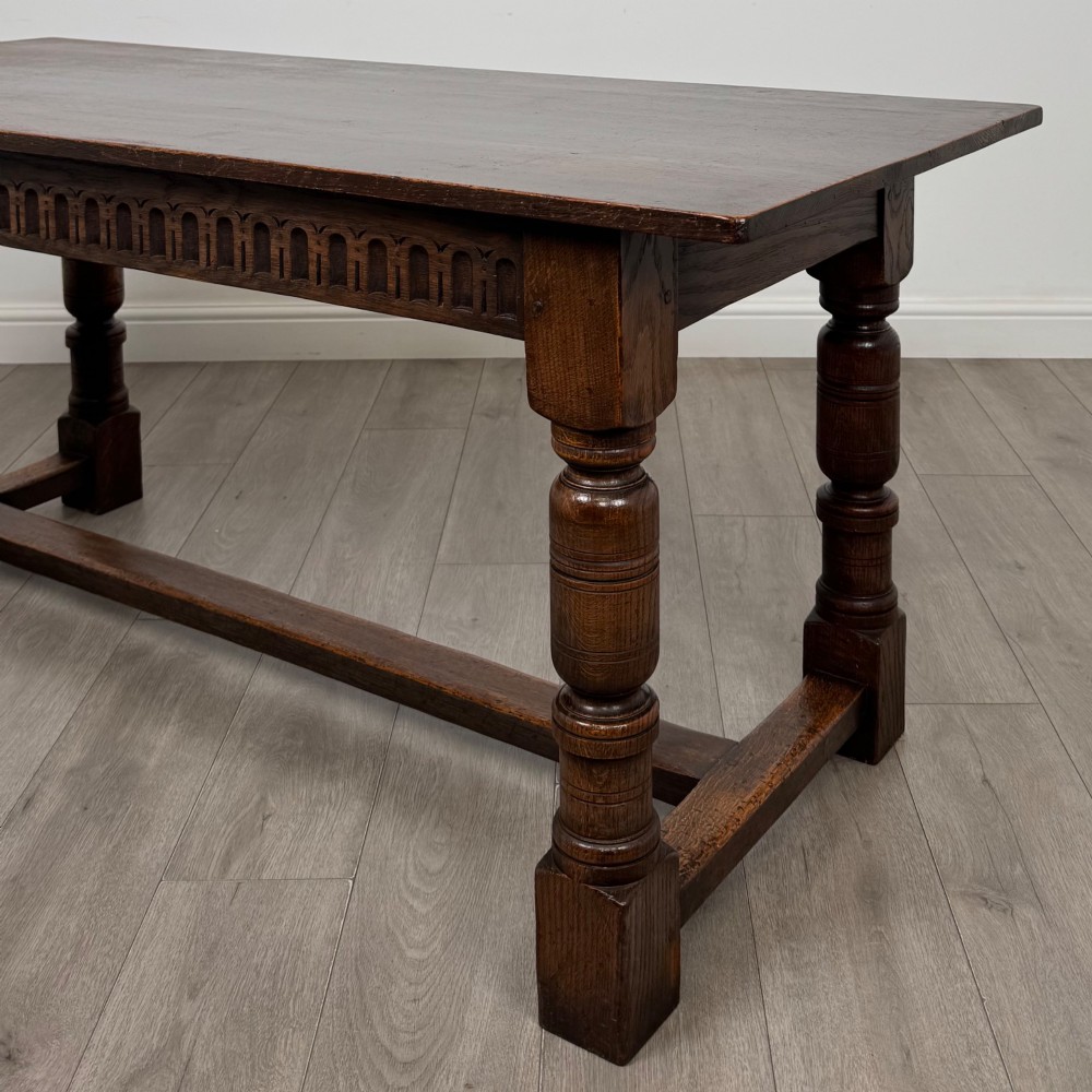 A dark oak rectangular refectory dining table featuring a carved arch frieze, four turned legs, and a low H-stretcher. View V