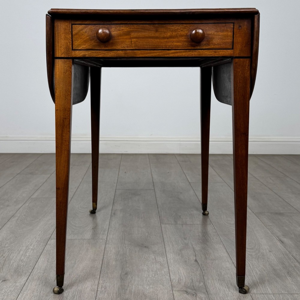A 19th-century Georgian mahogany Pembroke table featuring two hinged drop-leaf sides, a single drawer with wooden knobs, and square tapered legs on brass castors. View IV