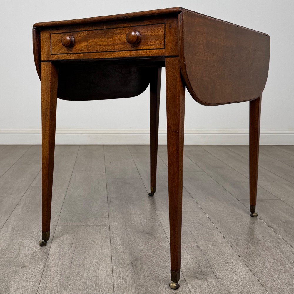 A 19th-century Georgian mahogany Pembroke table featuring two hinged drop-leaf sides, a single drawer with wooden knobs, and square tapered legs on brass castors. View VI
