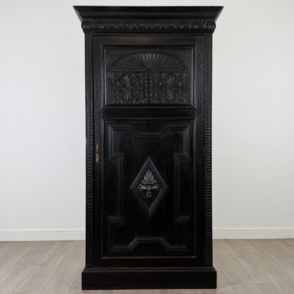 A single-door dark oak Victorian wardrobe featuring geometric panel mouldings, a carved relief face in a central diamond, and a pronounced decorative cornice.