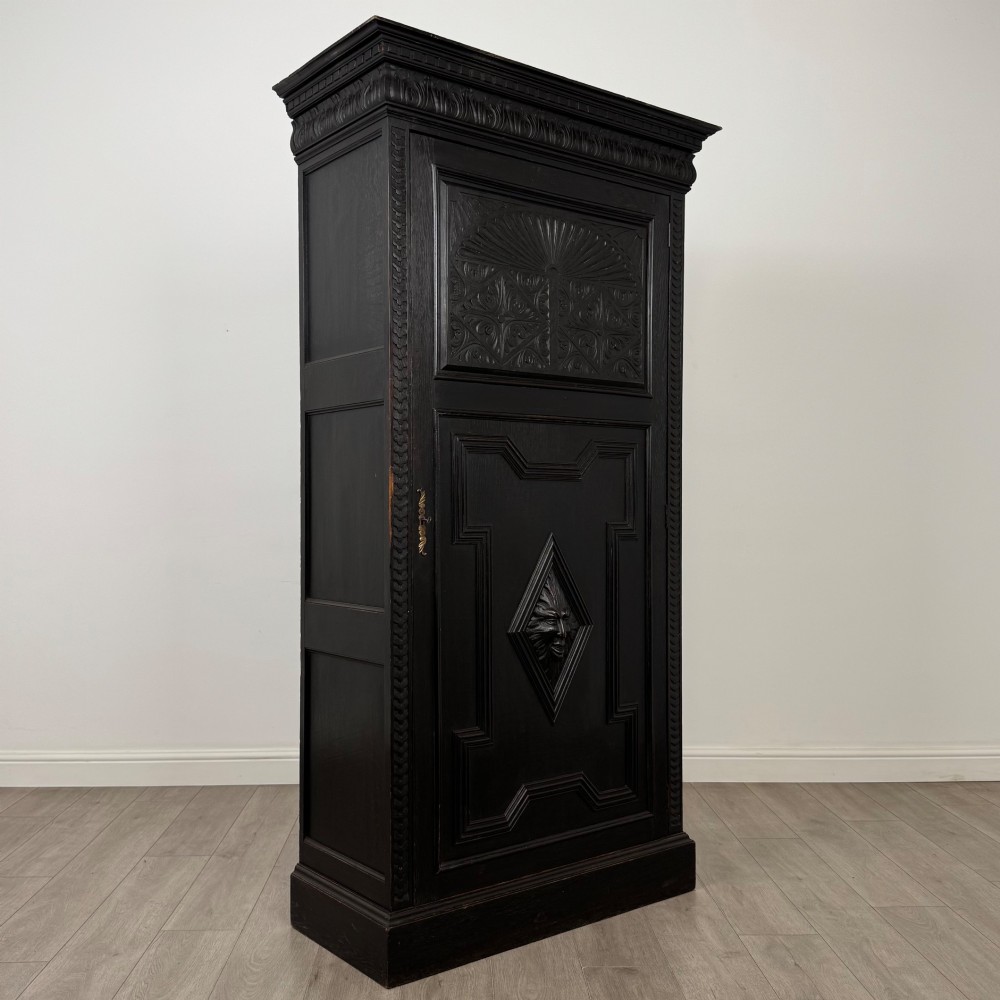 A single-door dark oak Victorian wardrobe featuring geometric panel mouldings, a carved relief face in a central diamond, and a pronounced decorative cornice. View IX