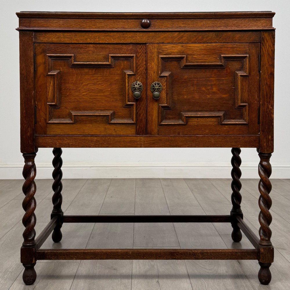 A dark oak Jacobean style cabinet featuring two doors with geometric fielded panels, twin brass drop handles, and four barley twist legs supported by low stretchers.