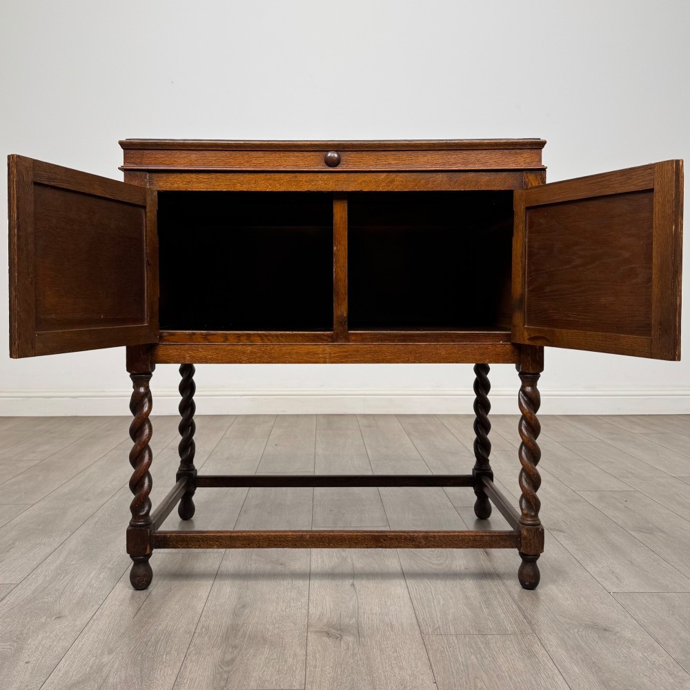 A dark oak Jacobean style cabinet featuring two doors with geometric fielded panels, twin brass drop handles, and four barley twist legs supported by low stretchers. View II