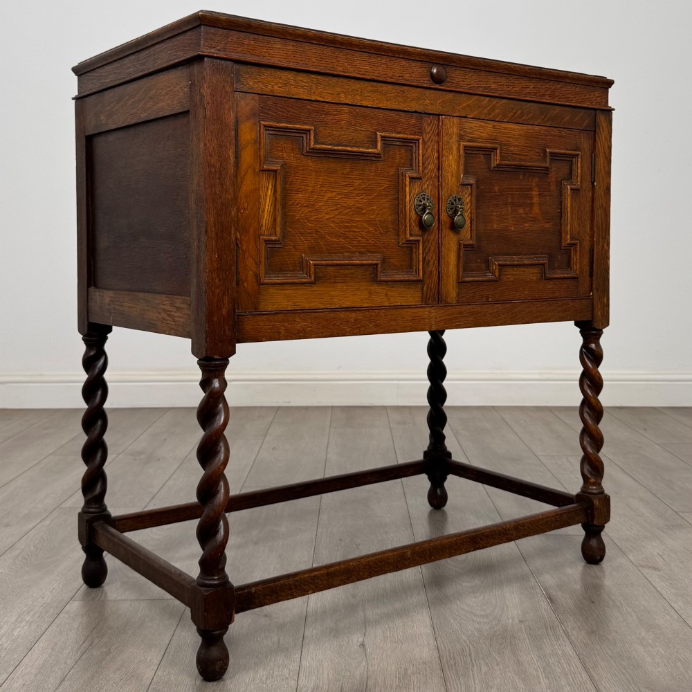 A dark oak Jacobean style cabinet featuring two doors with geometric fielded panels, twin brass drop handles, and four barley twist legs supported by low stretchers. View IX
