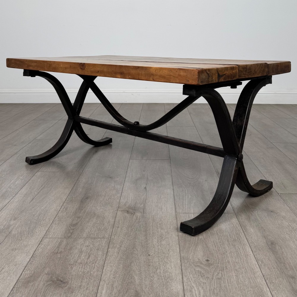 A low rectangular coffee table featuring a thick hardwood plank top set upon a dark curved iron X-frame base.