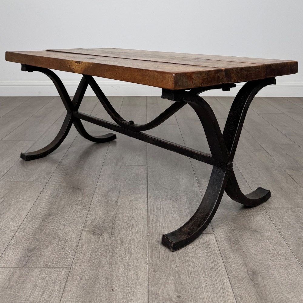 A low rectangular coffee table featuring a thick hardwood plank top set upon a dark curved iron X-frame base. View V