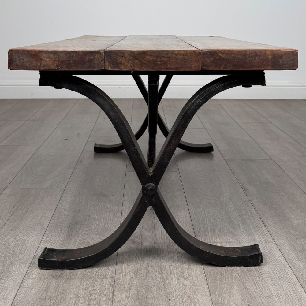 A low rectangular coffee table featuring a thick hardwood plank top set upon a dark curved iron X-frame base. View VI