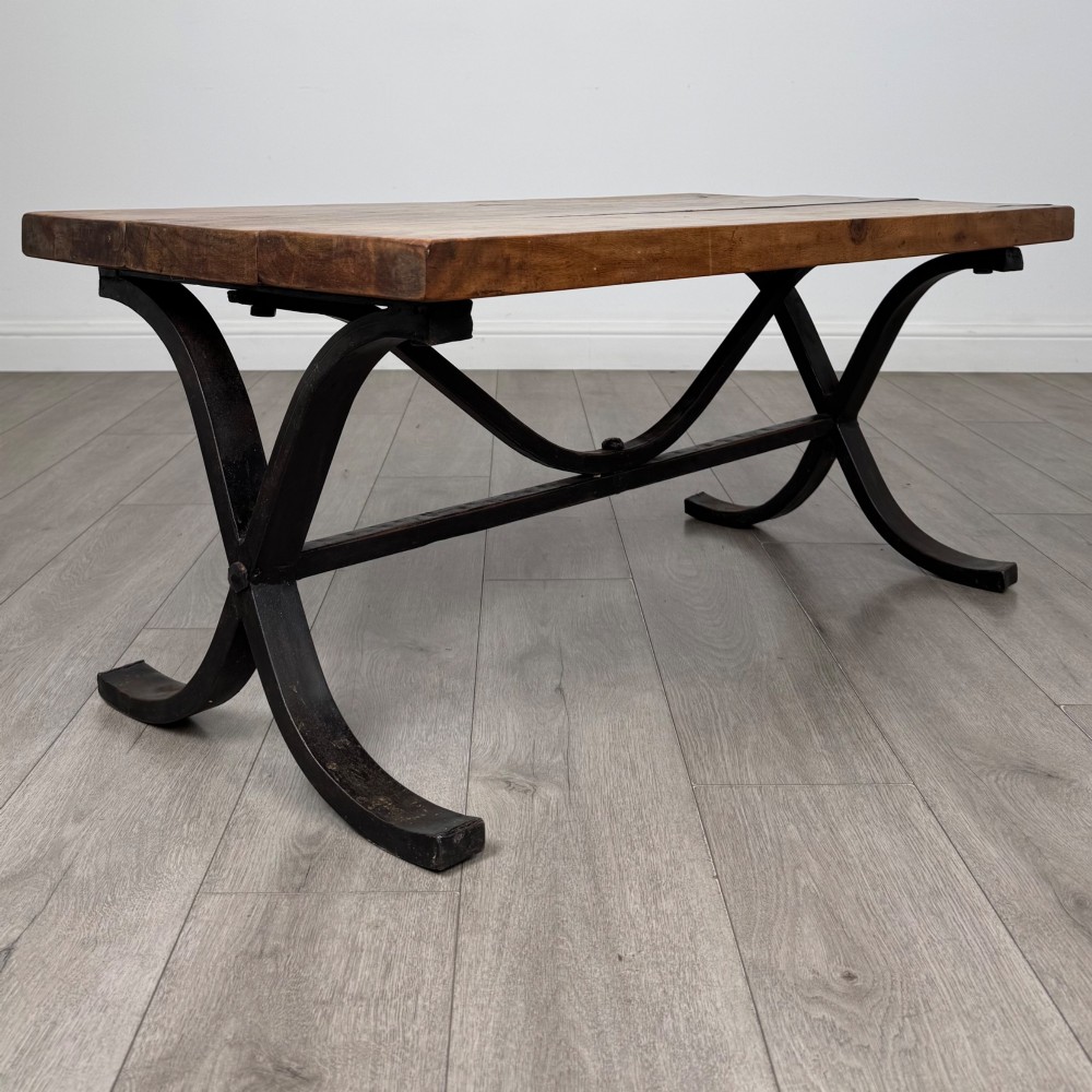 A low rectangular coffee table featuring a thick hardwood plank top set upon a dark curved iron X-frame base. View VII