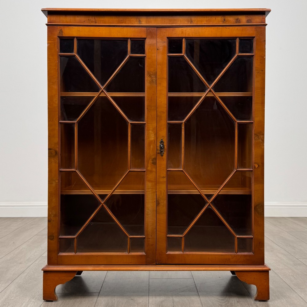 An antique yew wood display cabinet featuring two astragal glazed doors with geometric glazing bars, housing books on internal shelves, and resting on bracket feet. View II