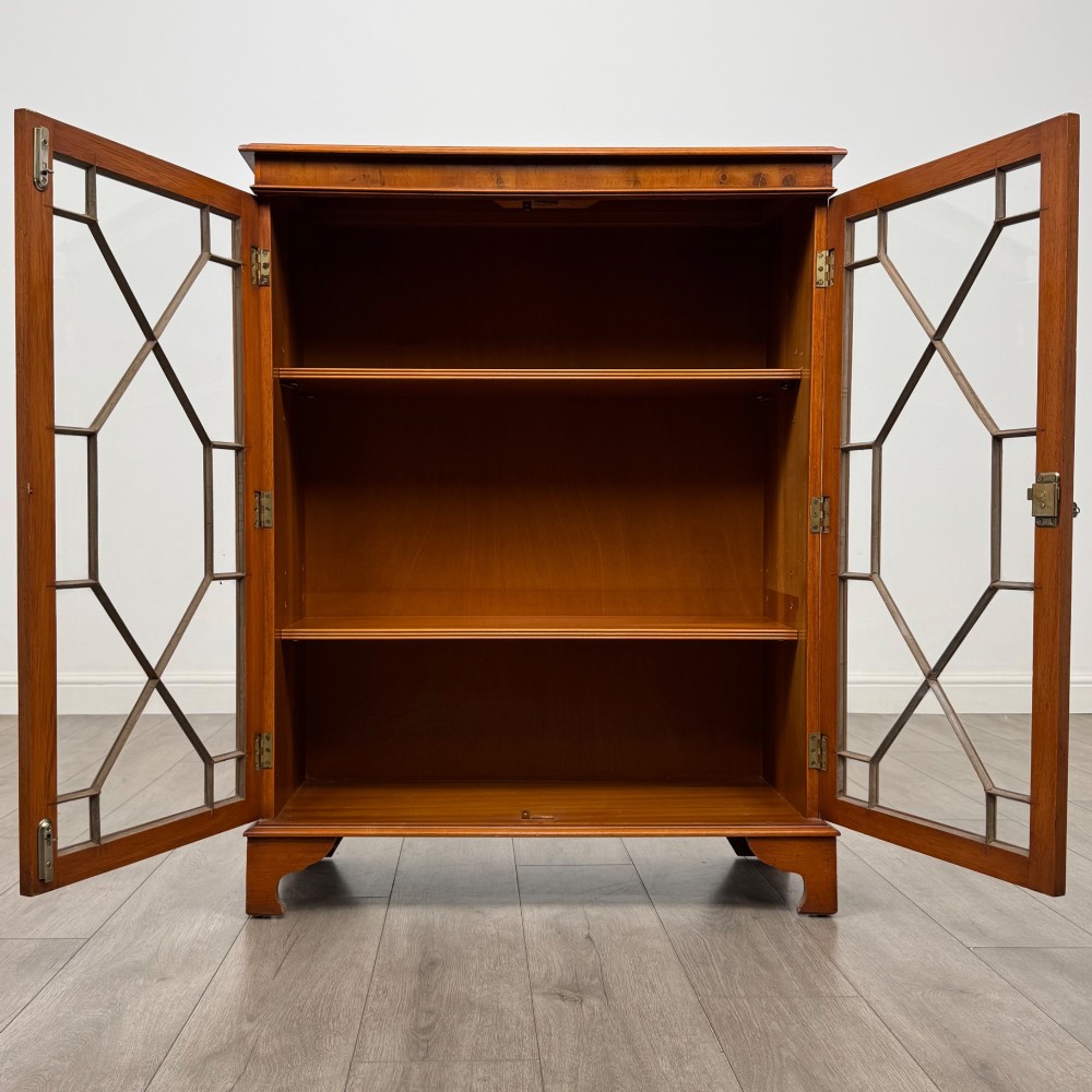 An antique yew wood display cabinet featuring two astragal glazed doors with geometric glazing bars, housing books on internal shelves, and resting on bracket feet. View III