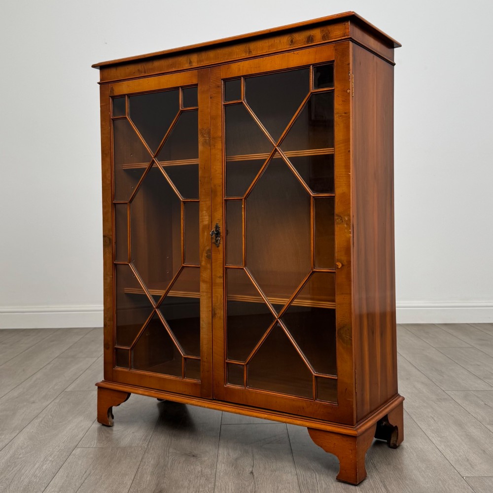 An antique yew wood display cabinet featuring two astragal glazed doors with geometric glazing bars, housing books on internal shelves, and resting on bracket feet. View IV