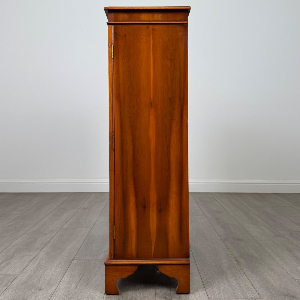 An antique yew wood display cabinet featuring two astragal glazed doors with geometric glazing bars, housing books on internal shelves, and resting on bracket feet. View V